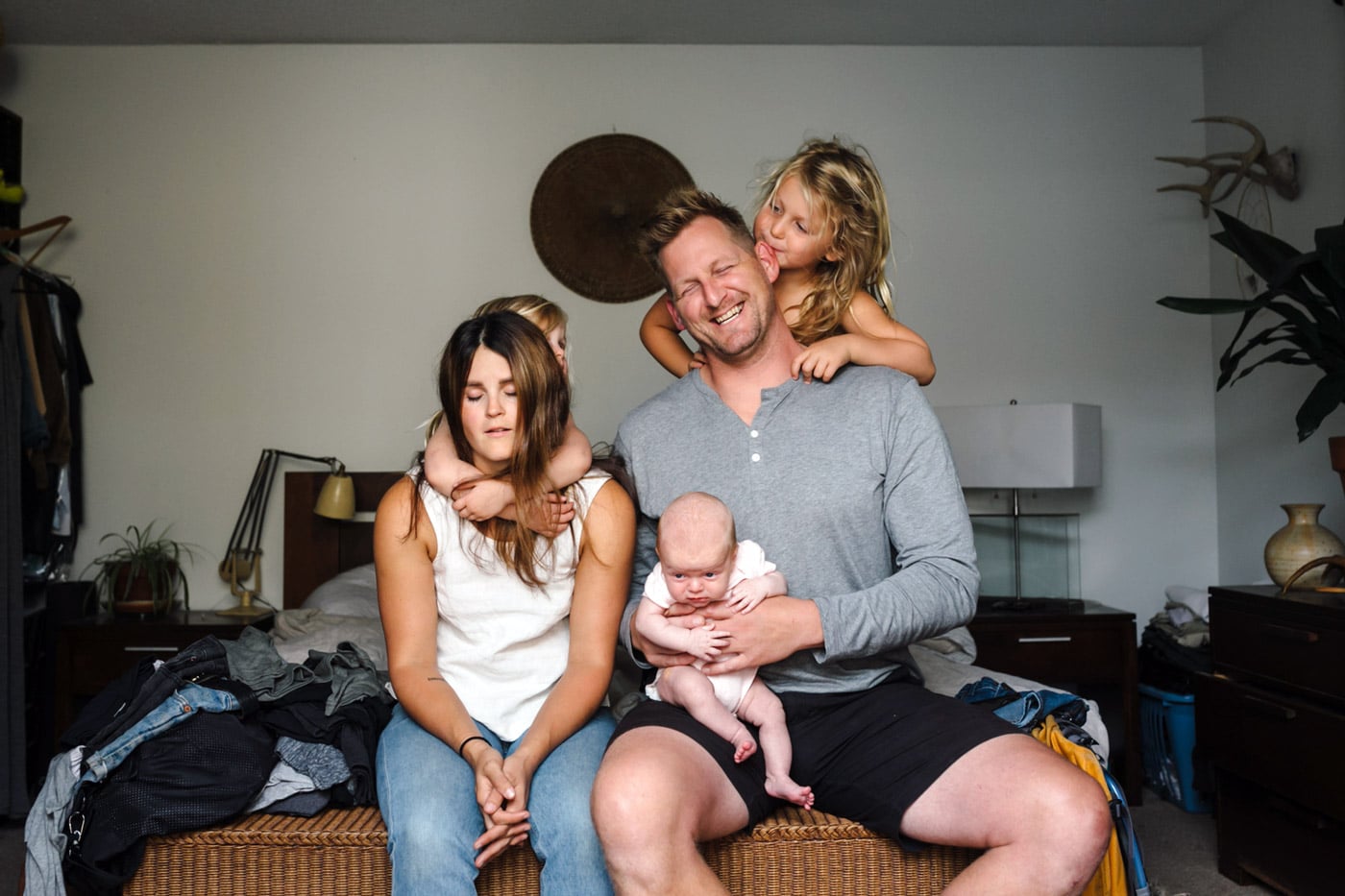 Familie på fem sitter på sengekanten, far og mor trøtte med barn på skuldre og fang